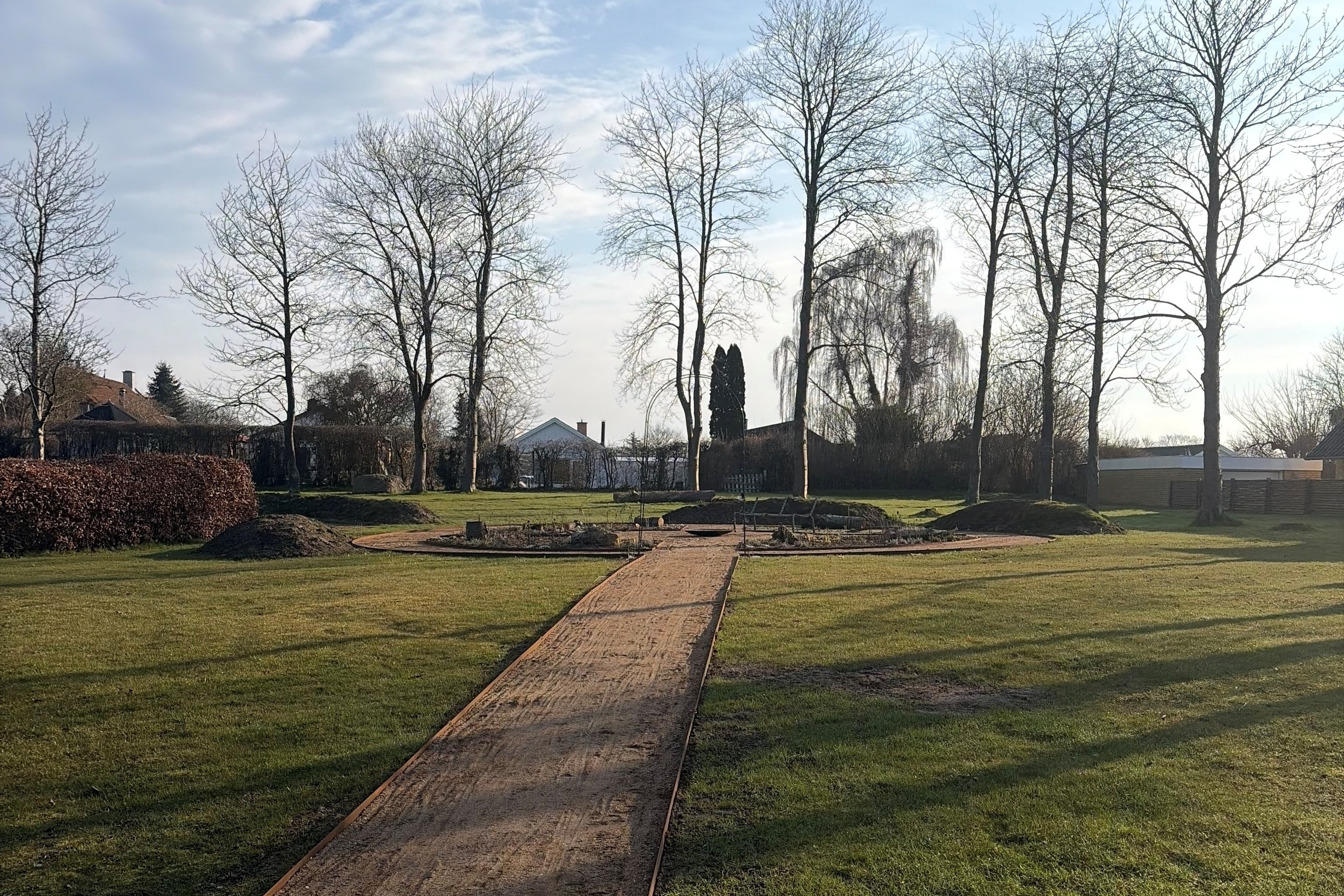 Foto af nyanlagte bede med planter og træstubbe midt på stor græspålæne. Høje træer i baggrunden